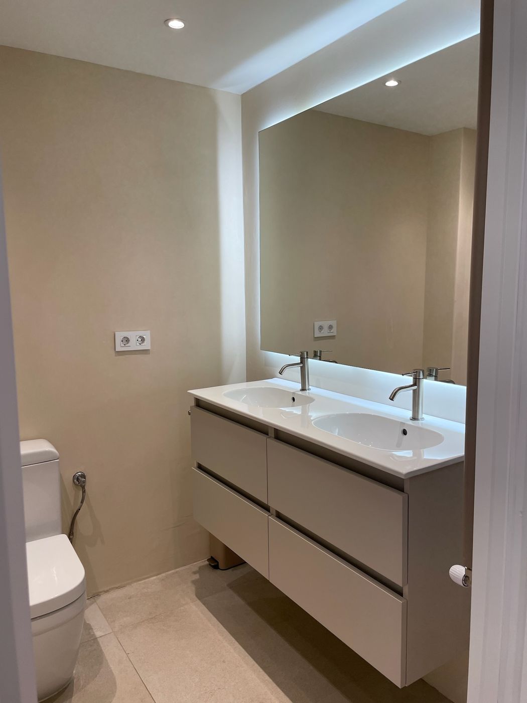 Baño reformado con doble lavabo, espejo iluminado y paredes en tono arena